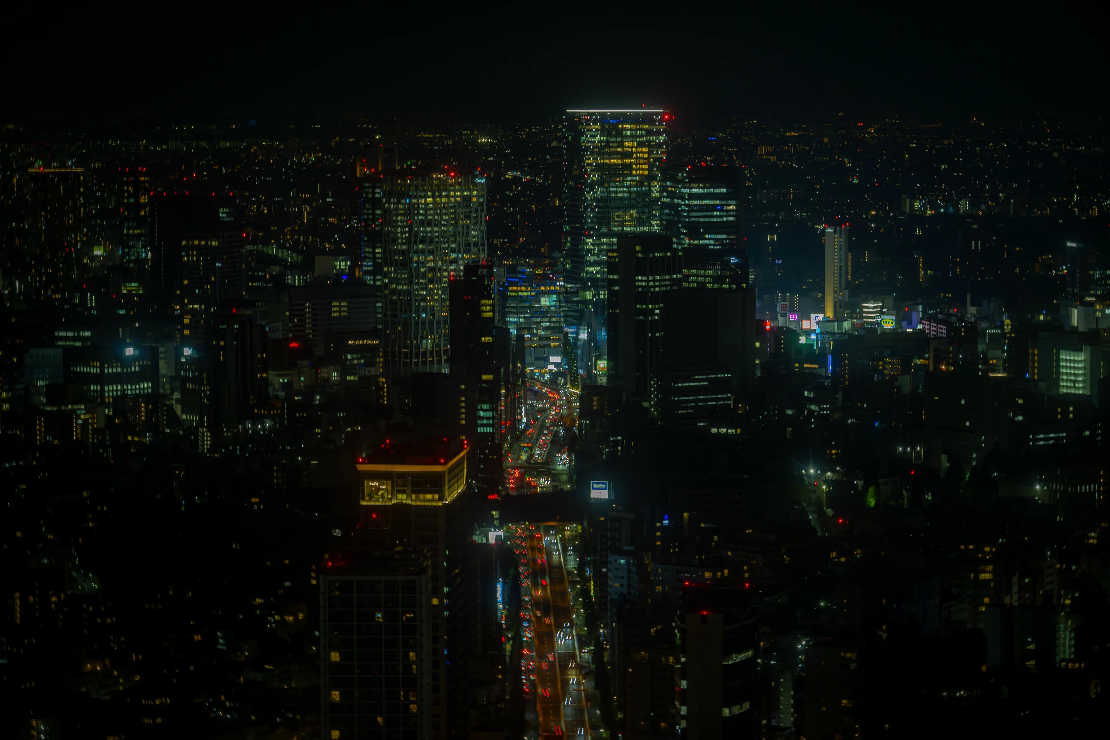 Tokyo at night from high above: dark blocks of buildings to the horizon, a cluster of lit towers at the top and a single avenue of red and white traffic running up through the middle.