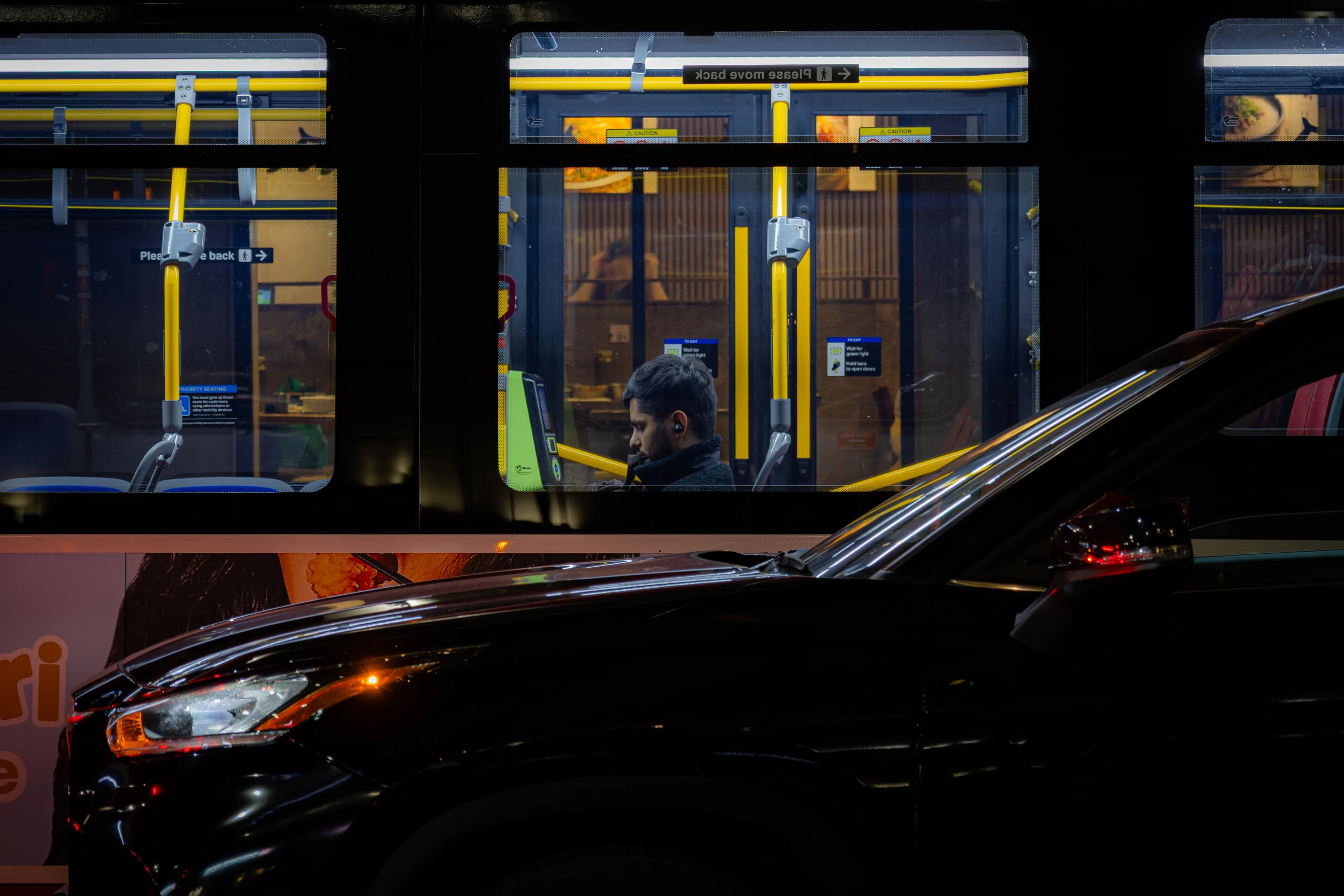 A rider lit up in a transit window at night, yellow poles and a 'Please move back' sign around them, the black hood of a passing car cutting across the lower half of the frame.