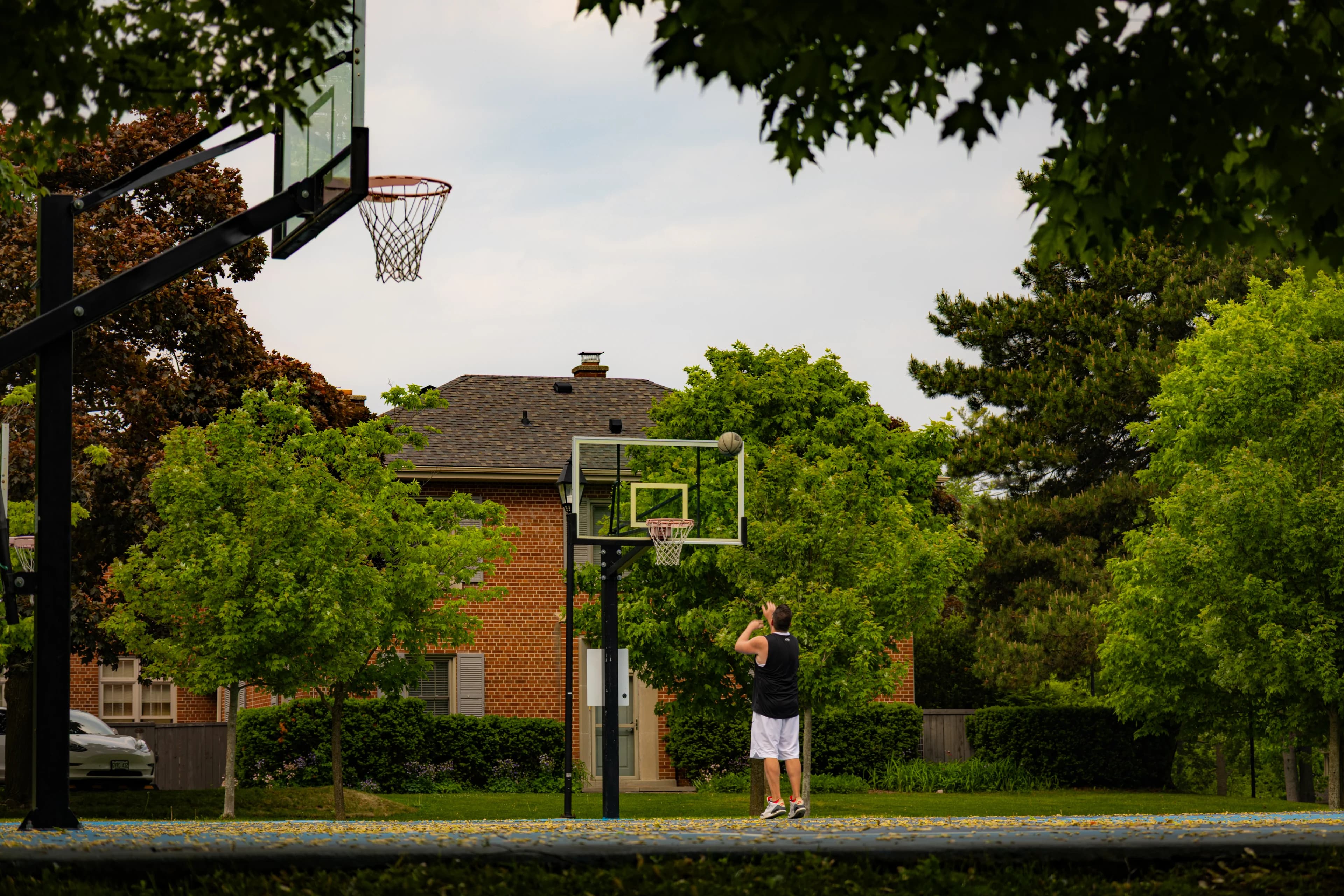 A lone player shooting at an outdoor hoop on a grey June afternoon, the ball at the backboard, a red-brick house and green trees behind the court.