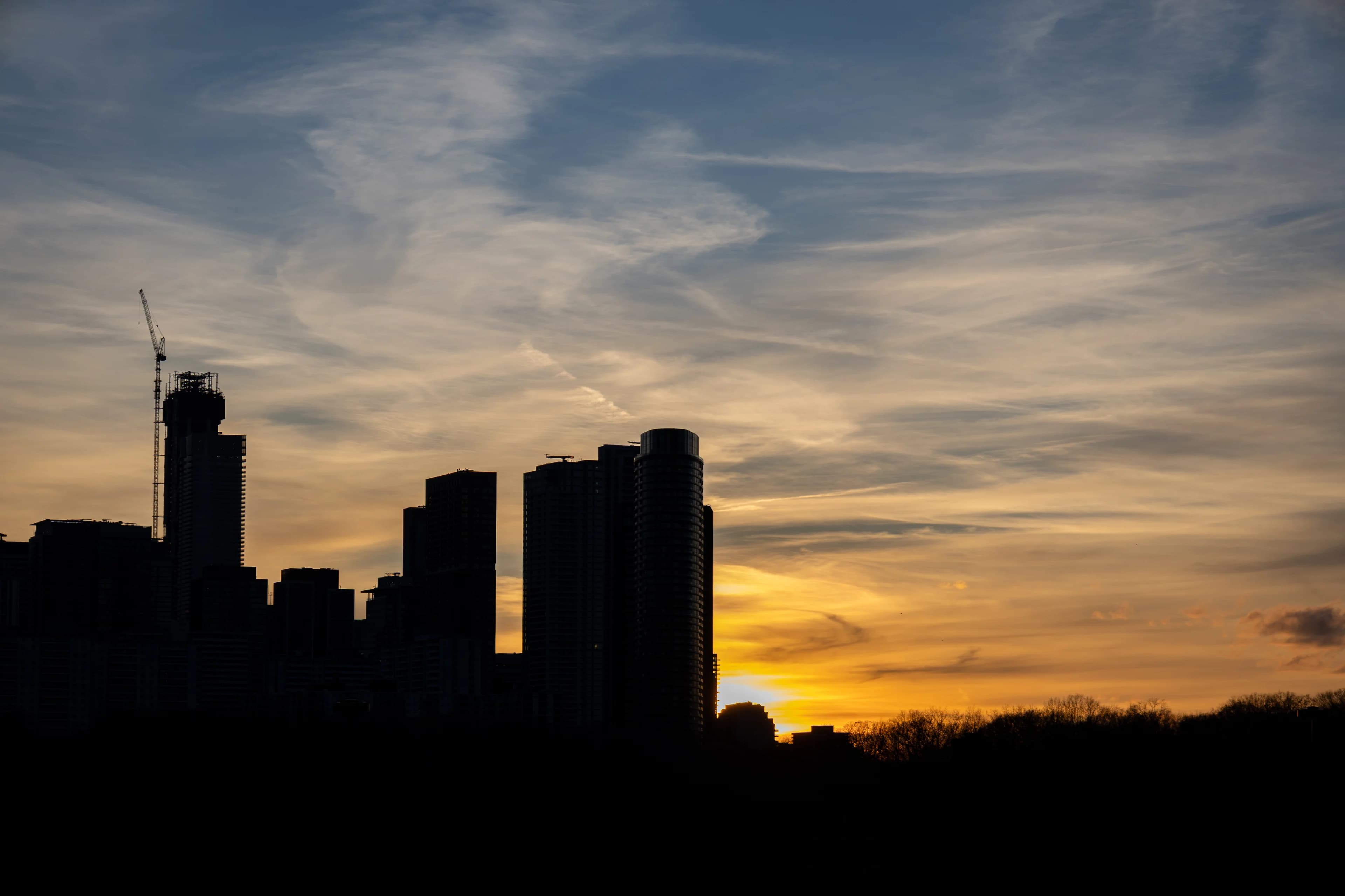 Condo towers and a construction crane in black silhouette against an orange sunset streaked with cloud, the sun just under the rooftops on the left.