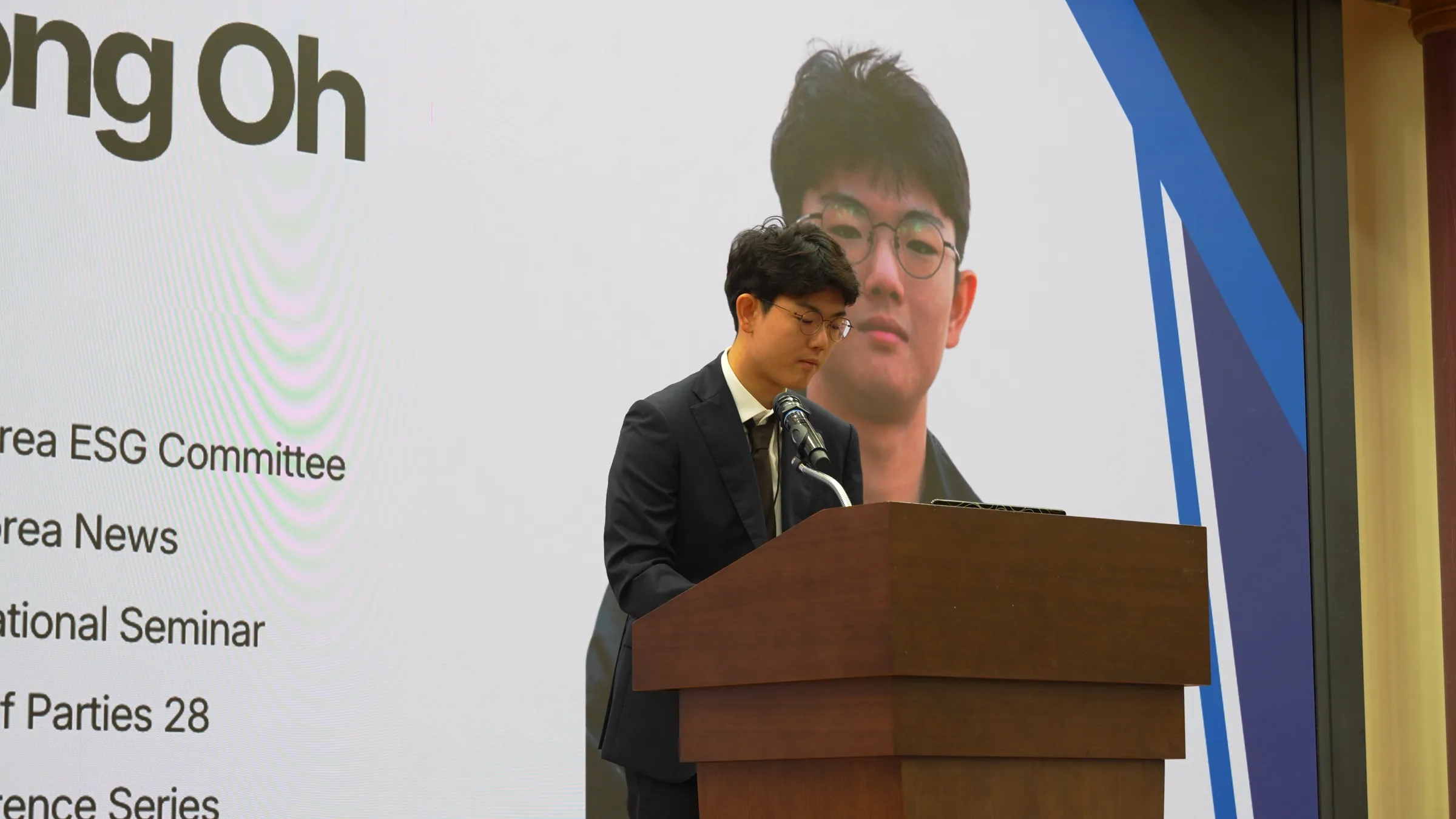 A young man in glasses and a dark suit reads from a wooden lectern with a microphone, in front of a large screen showing his own portrait and a list of roles including Korea ESG Committee.