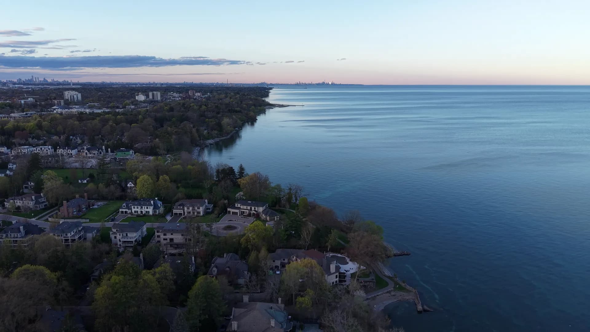 Blue-hour aerial view of a wooded shoreline curving along a calm lake, a distant skyline under a pink band of sky.