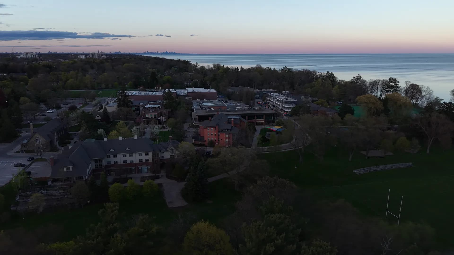 Dusk aerial view of red-brick and stone school buildings among trees, the lake and a pink horizon beyond.