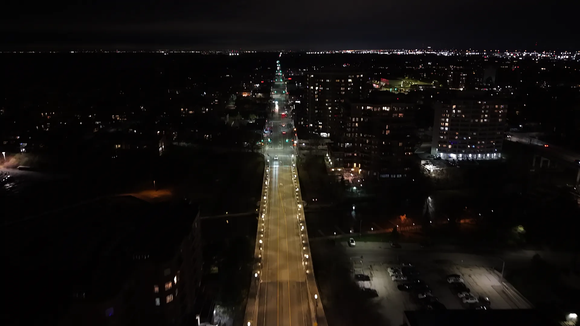 Night drone view down a lit avenue running to the horizon, apartment blocks glowing on either side.