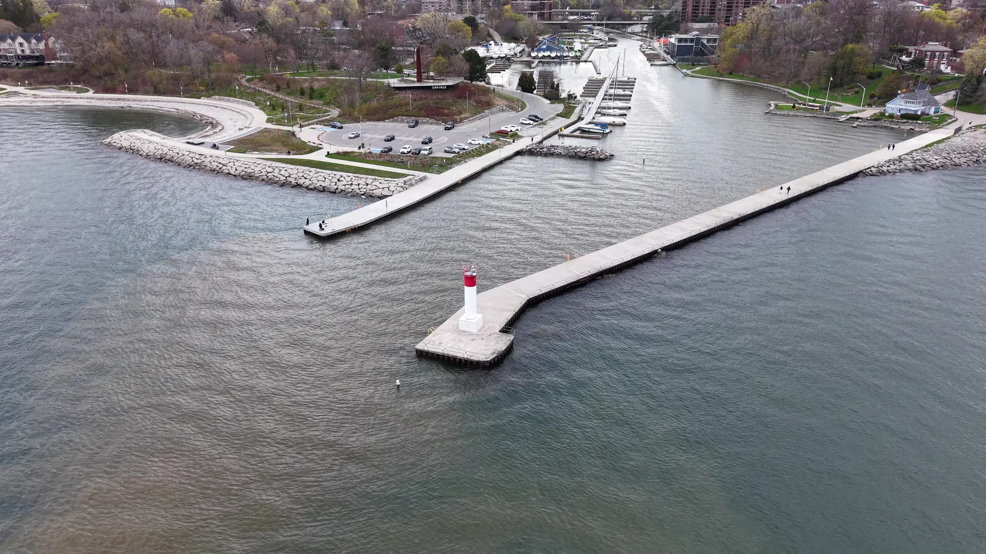 Aerial view of a long concrete pier ending in a small red-and-white lighthouse, brown creek water spreading into grey lake water.