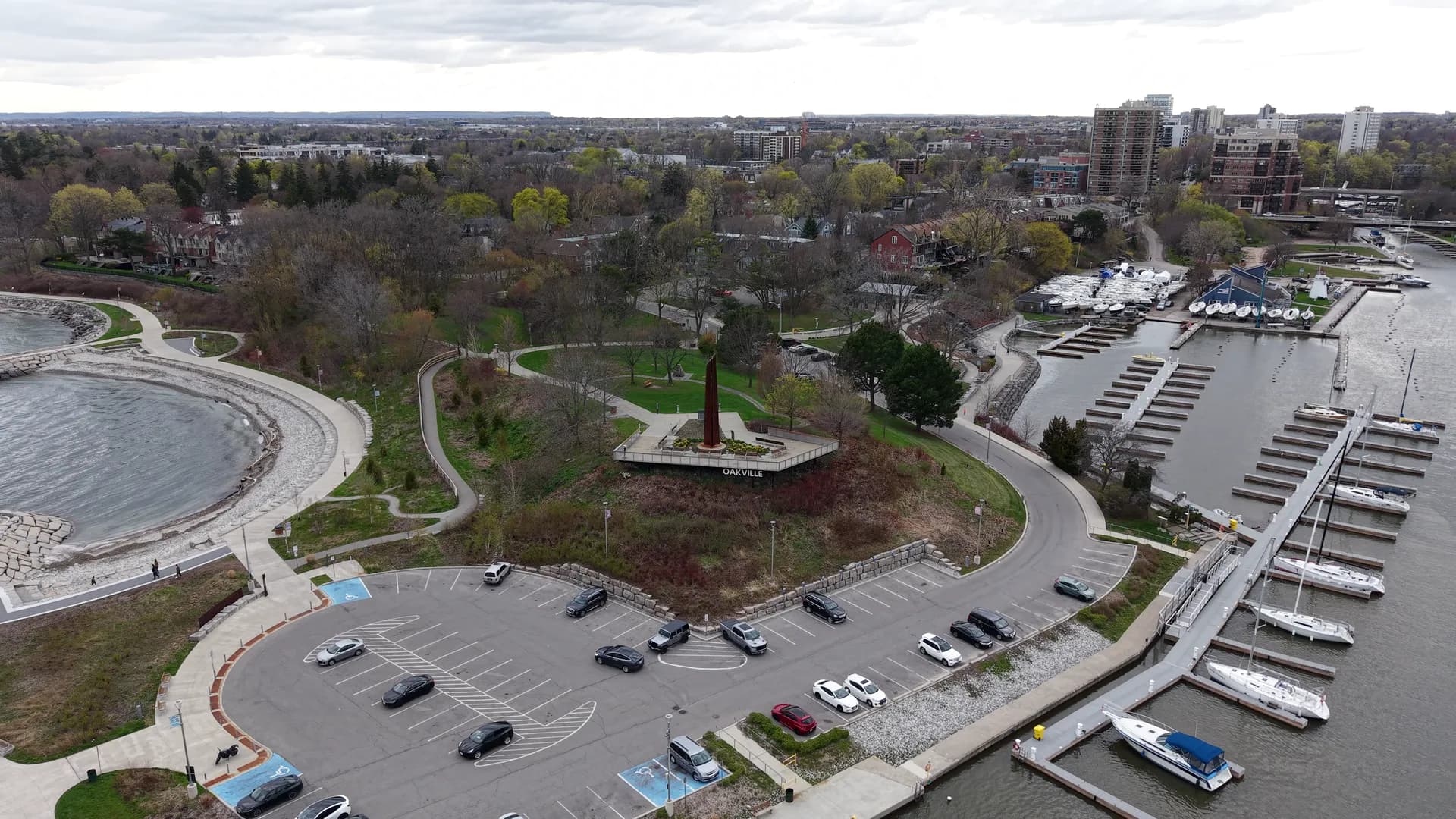 Aerial view of a lakeside park with a tall rust-coloured sculpture on a raised terrace, a curving breakwater and rows of boat slips.