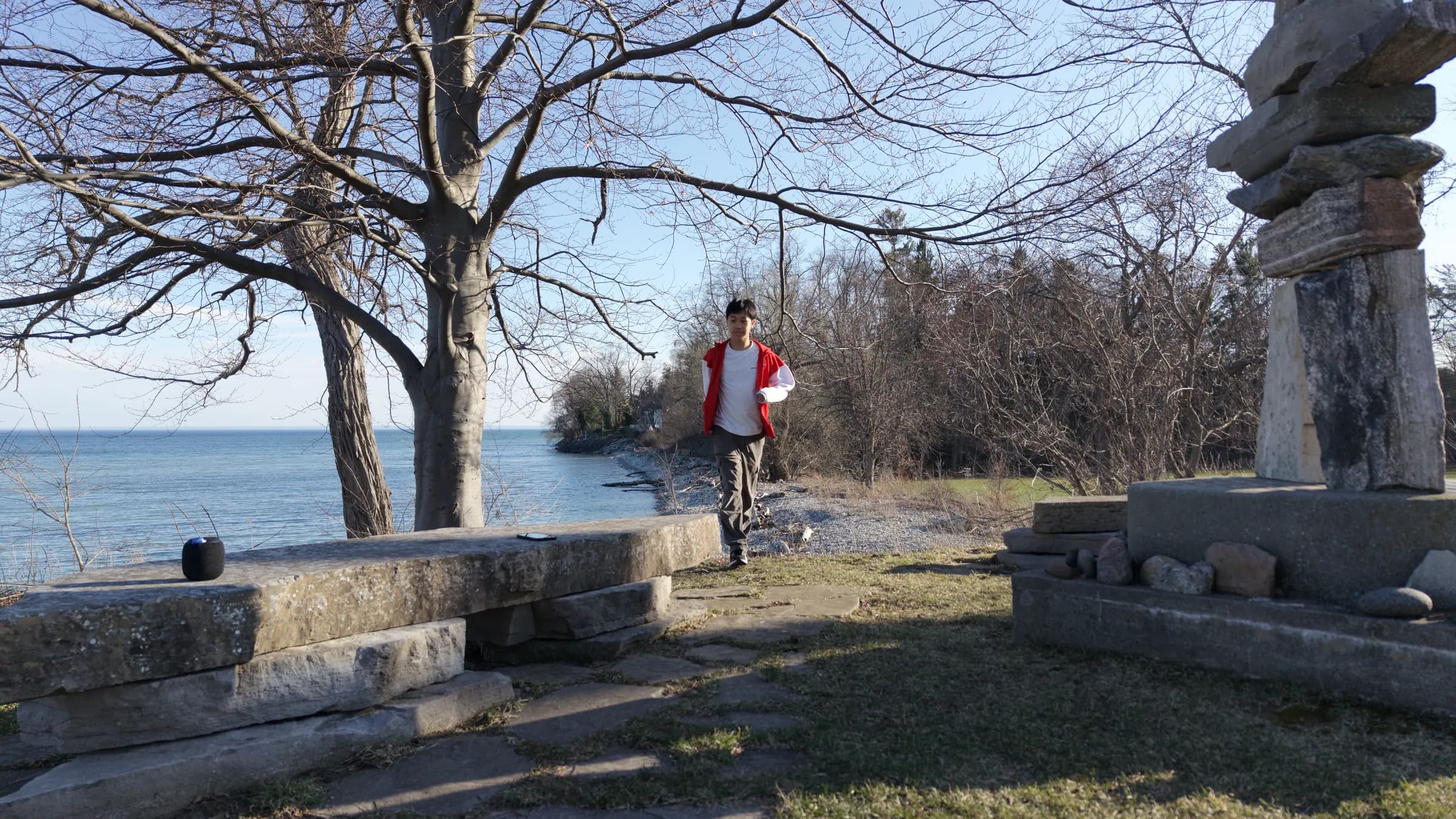 A person in a red jacket walks along a lakeside lookout beside a stone inukshuk and bare trees, with Lake Ontario stretching to the horizon.