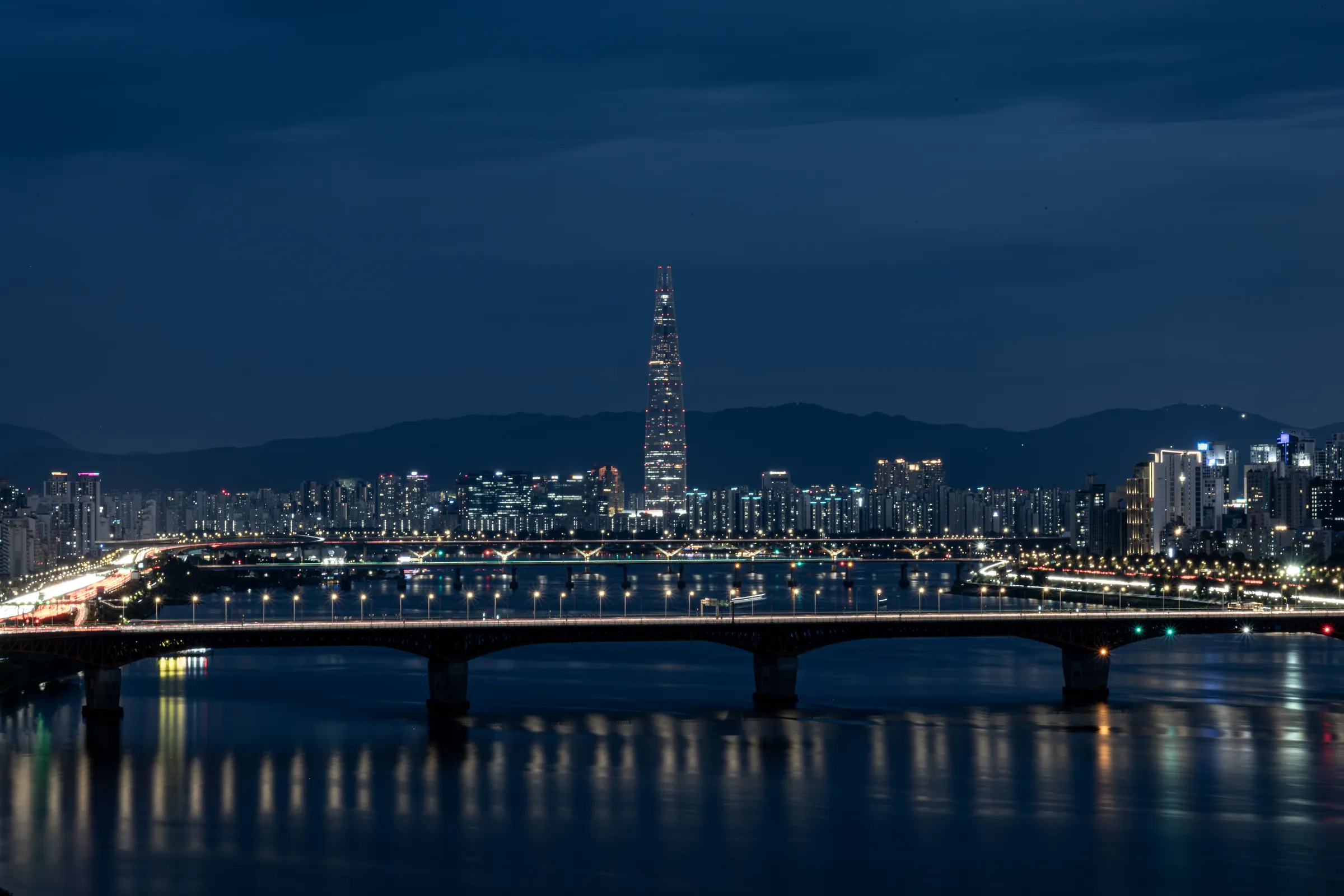 Night view across the Han River to a lit bridge and the city skyline, with Lotte World Tower rising in the distance under a deep blue sky.