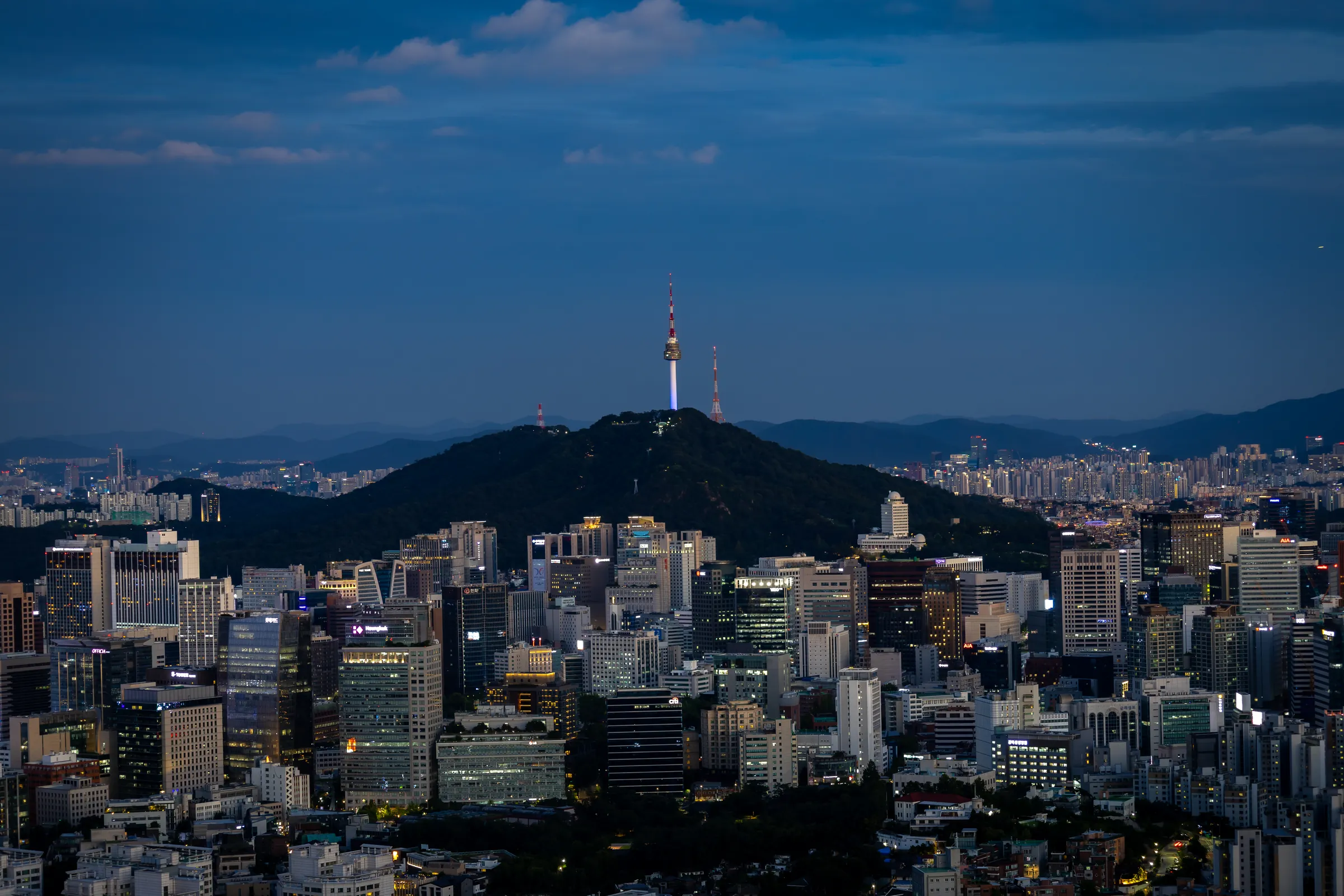 Dense Seoul cityscape at dusk with Namsan and N Seoul Tower rising on the hill behind office towers.