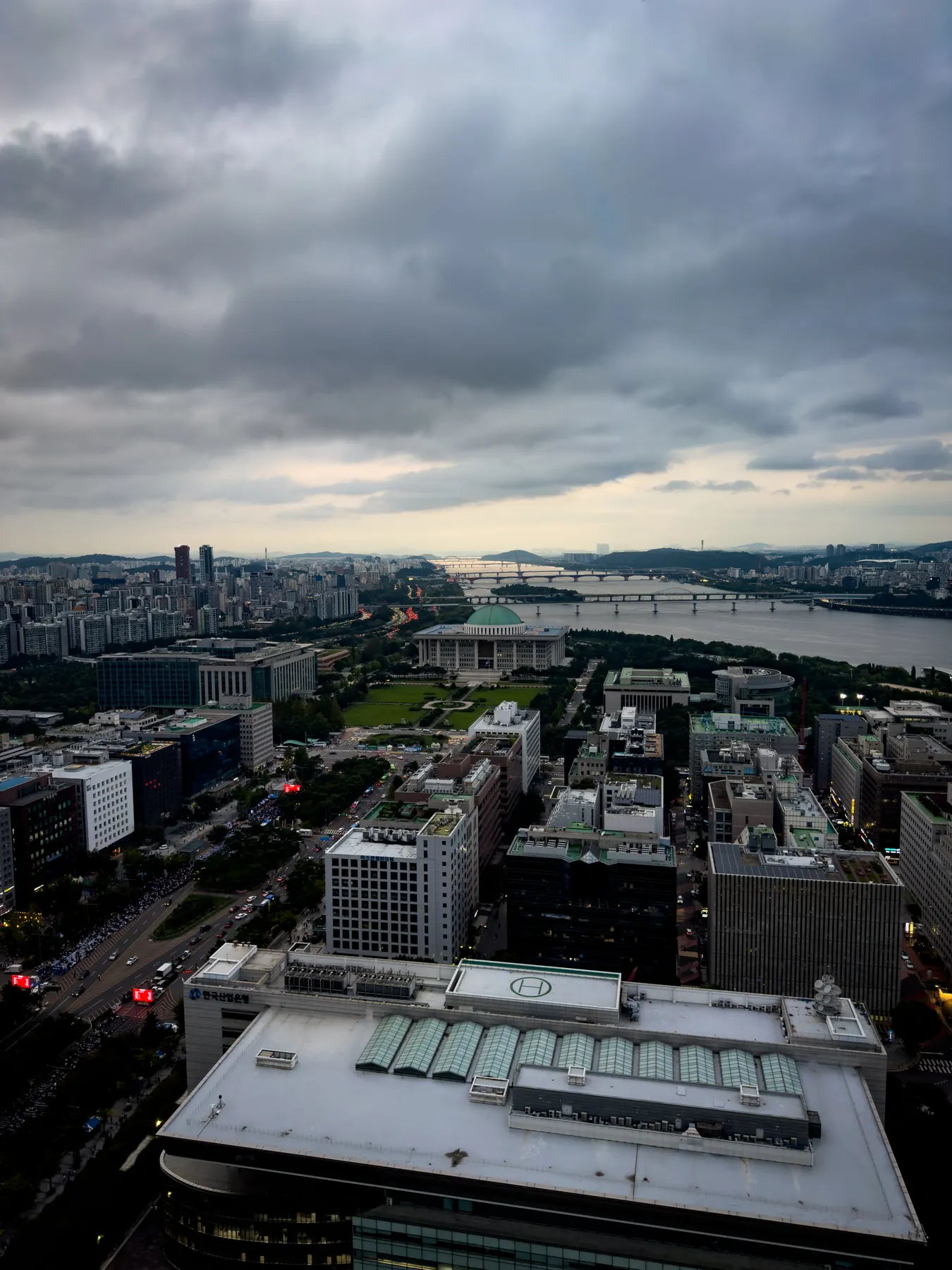 High aerial view over Yeouido under heavy clouds: office rooftops in the foreground, the green-domed National Assembly building and its lawn beyond, and the Han River with its bridges to the horizon.