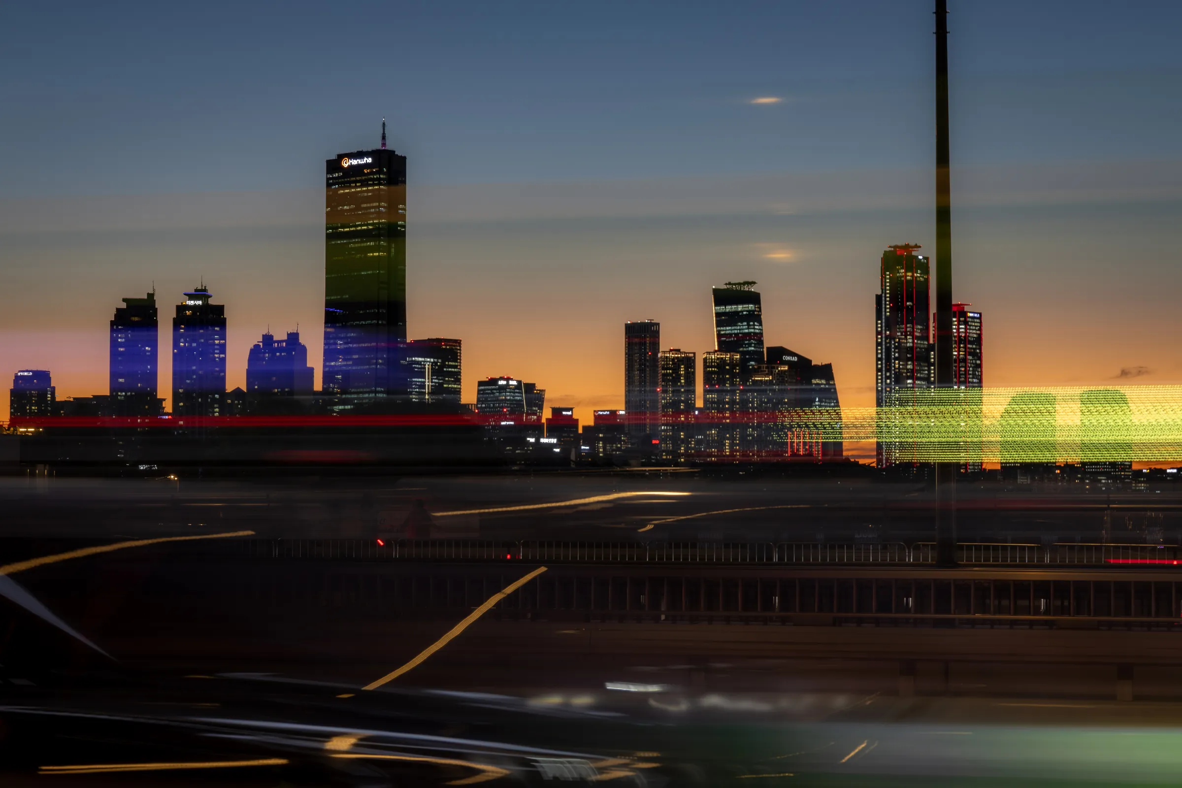 Yeouido towers, including the gold-glass 63 Building, silhouetted against an orange dusk sky, with blurred traffic light trails streaking across the foreground.