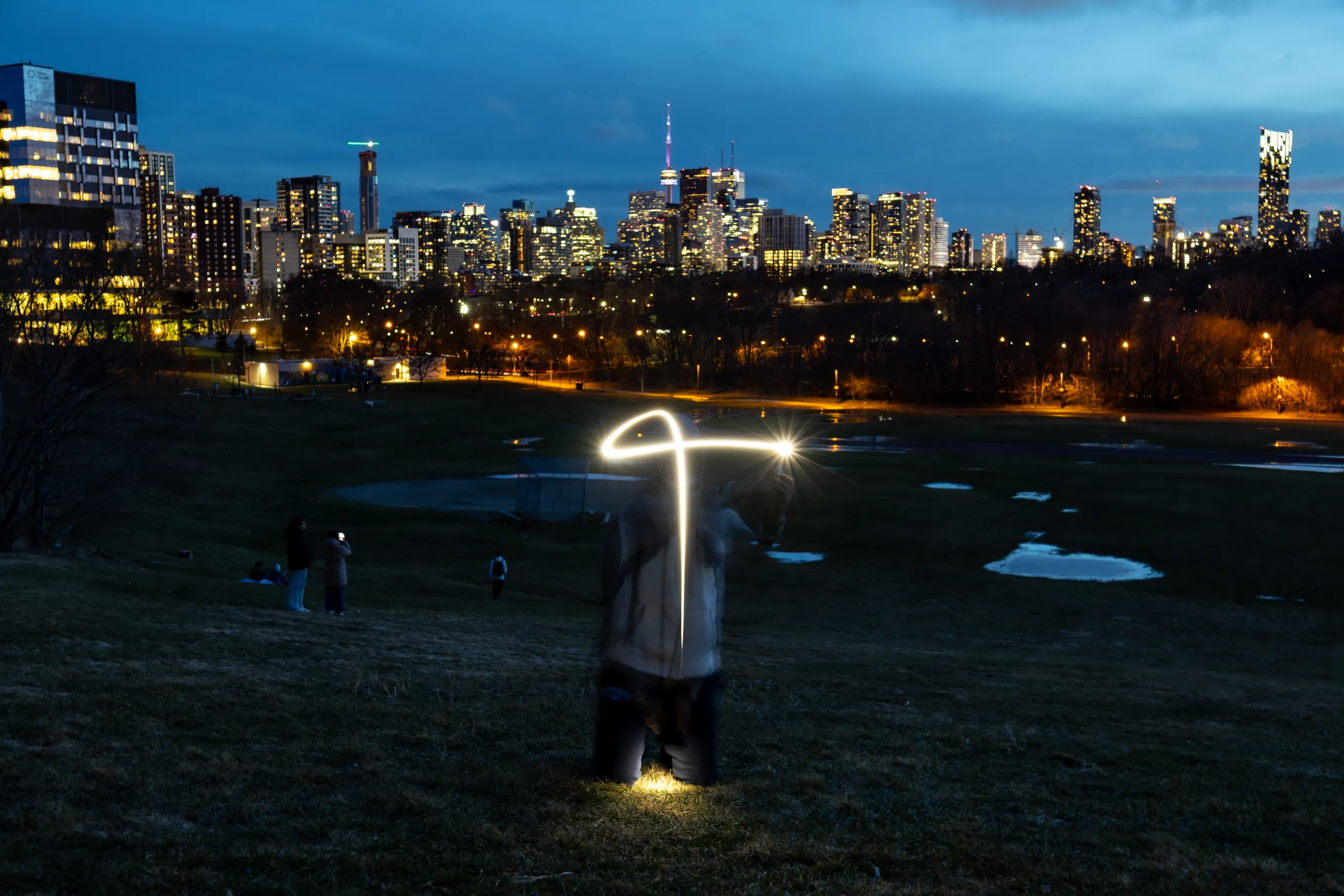 A long-exposure light painting glows on a dark park slope, with the lit Toronto skyline across the horizon at blue hour.