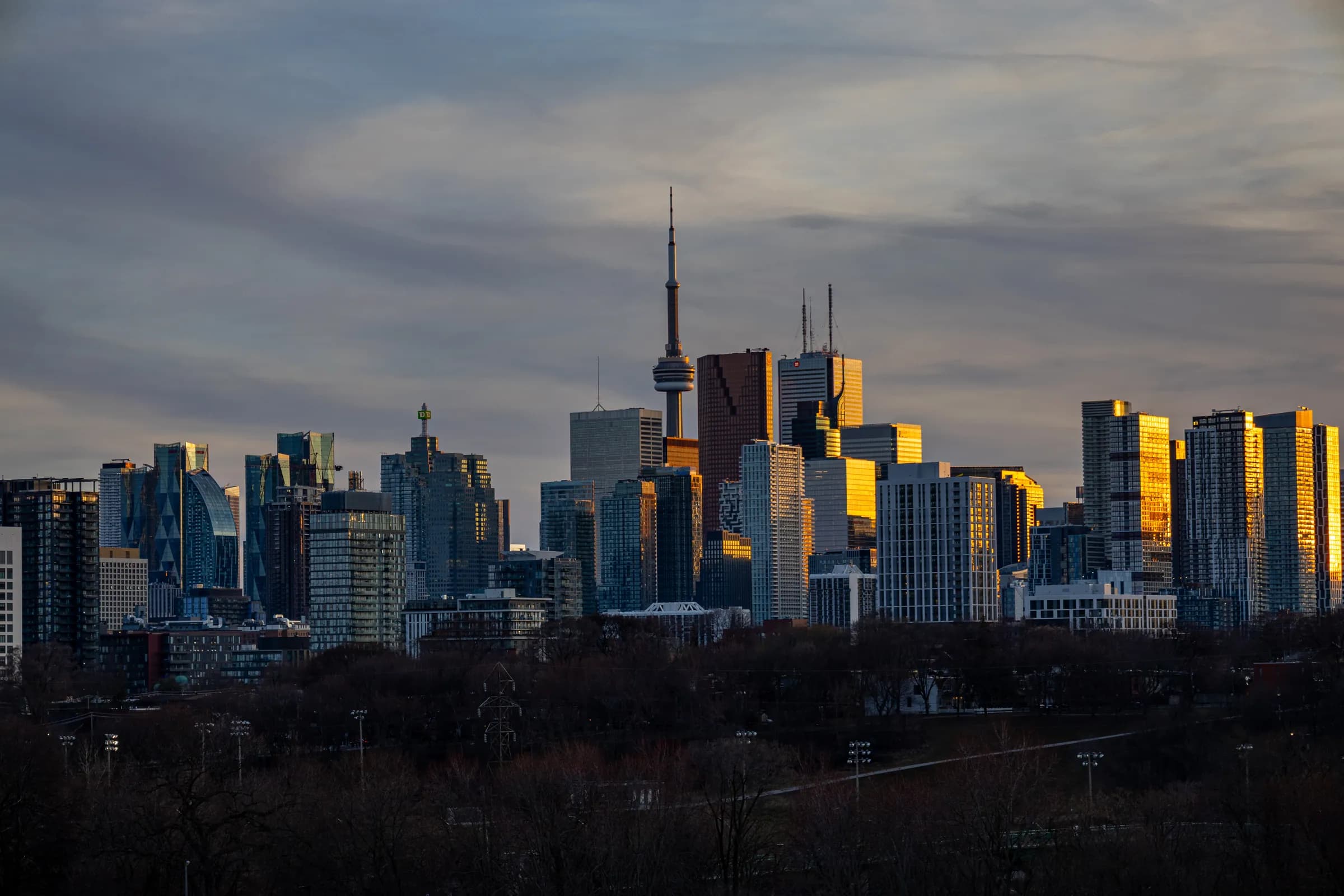 Downtown Toronto towers and the CN Tower catch low golden sun above a dark band of trees, under a cloudy sky.