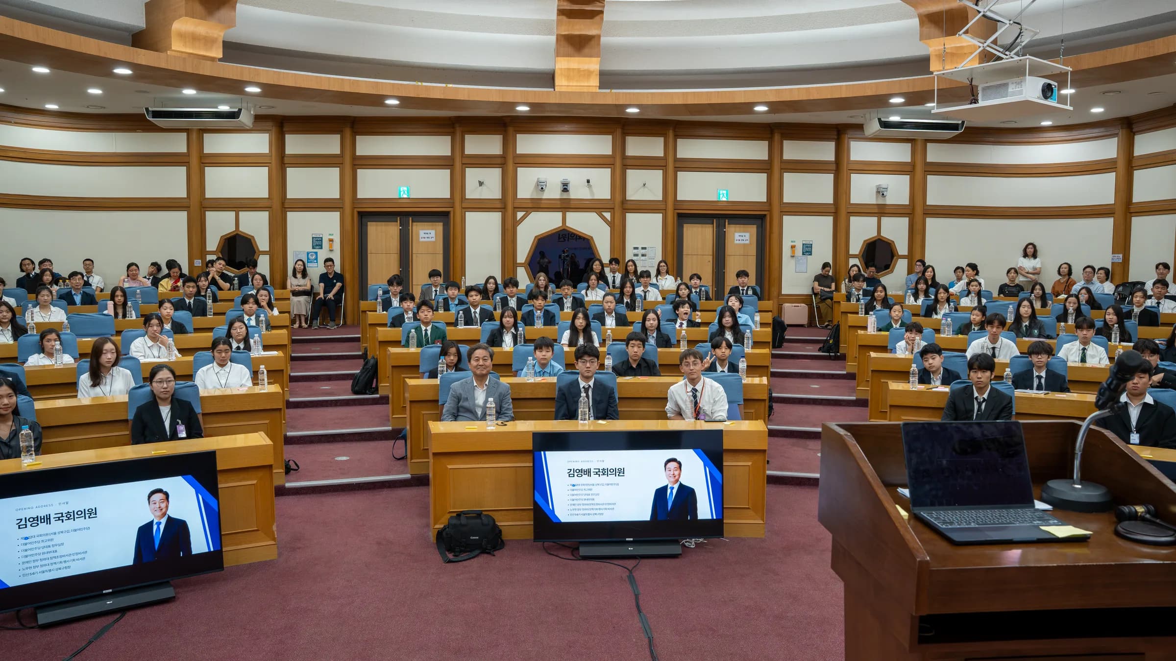 Delegates in suits and school uniforms fill the curved wooden desks of a National Assembly conference hall, facing screens that show Assemblyman Kim Young-bae.