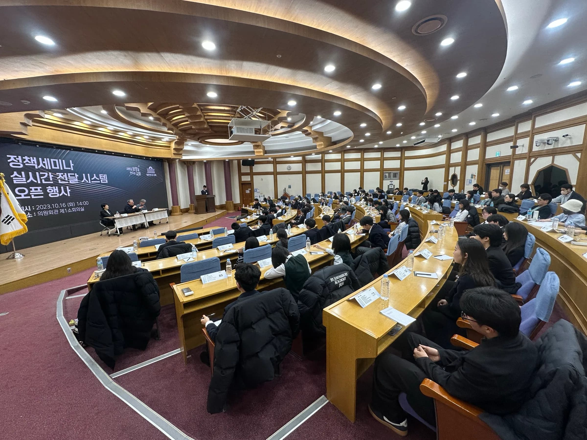 A curved National Assembly conference hall under a tiered wooden ceiling, full of students at long desks facing a panel on stage beside the Korean flag.