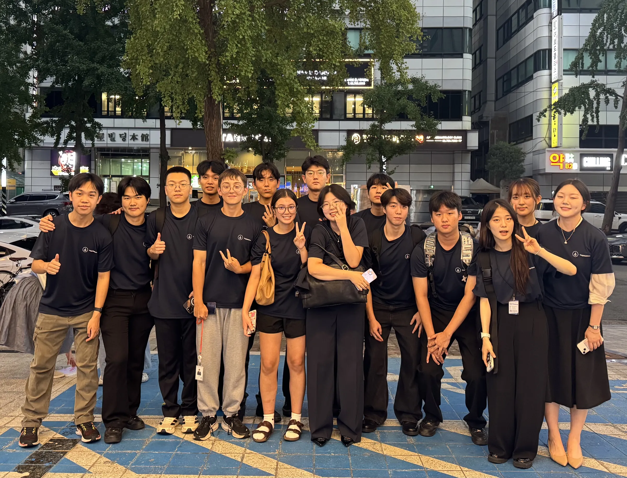 About sixteen young team members in matching navy Vegaplatform T-shirts stand together on a city pavement at dusk, some giving peace signs and thumbs-up.